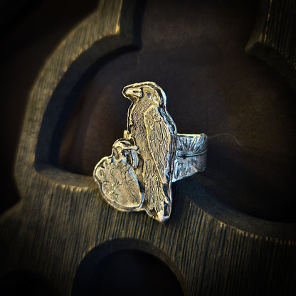 Sterling silver raven and human heart ring with a feather band on a dark wood background