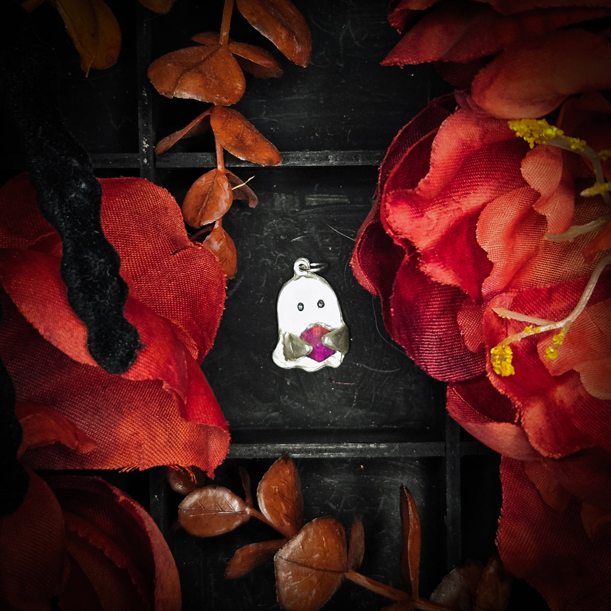 A small sterling silver ghost-shaped pendant with a gem, centered on a dark textured background surrounded by red fabric flowers.