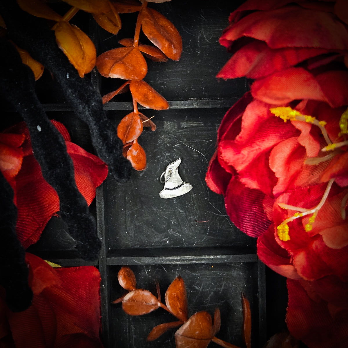 A small sterling silver witches hat charm, displayed on a dark textured background surrounded by red and orange autumn leaves and flowers.