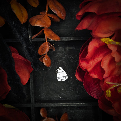 A small sterling silver candy corn charm, displayed on a dark textured background surrounded by red and orange autumn leaves and flowers.