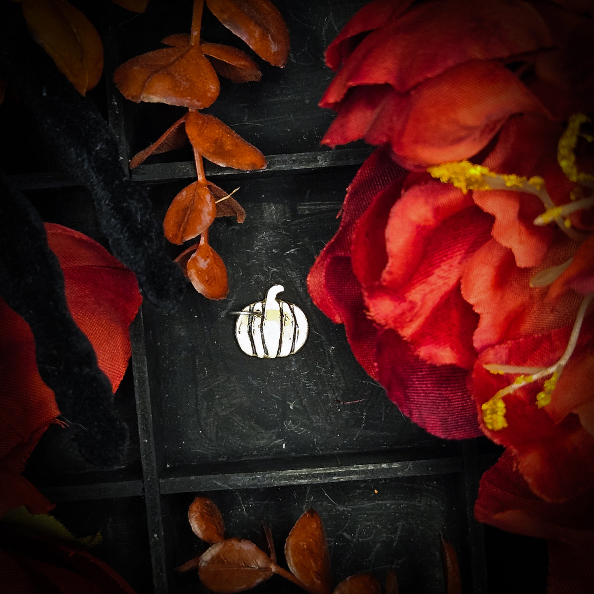 A small brass pumpkin charm, displayed on a dark textured background surrounded by red and orange autumn leaves and flowers.