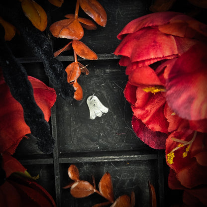 A small sterling silver floating ghost charm, displayed on a dark textured background surrounded by red and orange autumn leaves and flowers.