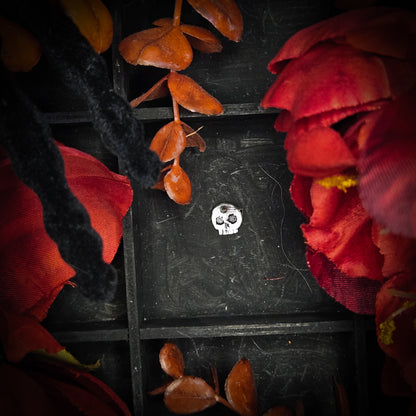 A small sterling silver mini skull charm, displayed on a dark textured background surrounded by red and orange autumn leaves and flowers.