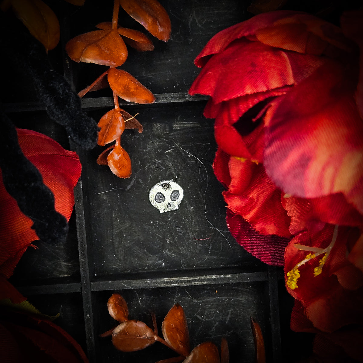 A small sterling silver skull charm, displayed on a dark textured background surrounded by red and orange autumn leaves and flowers.