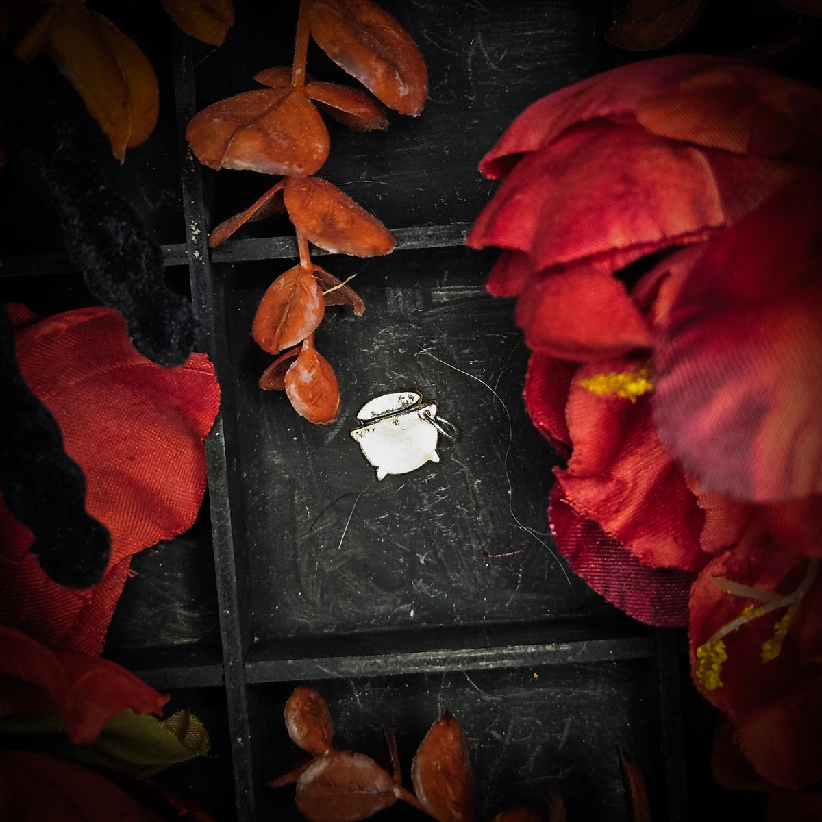 A small brass cauldron charm, displayed on a dark textured background surrounded by red and orange autumn leaves and flowers.