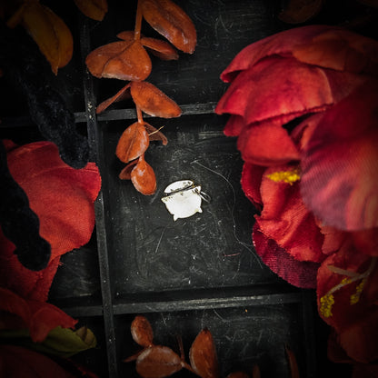 A small brass cauldron charm, displayed on a dark textured background surrounded by red and orange autumn leaves and flowers.
