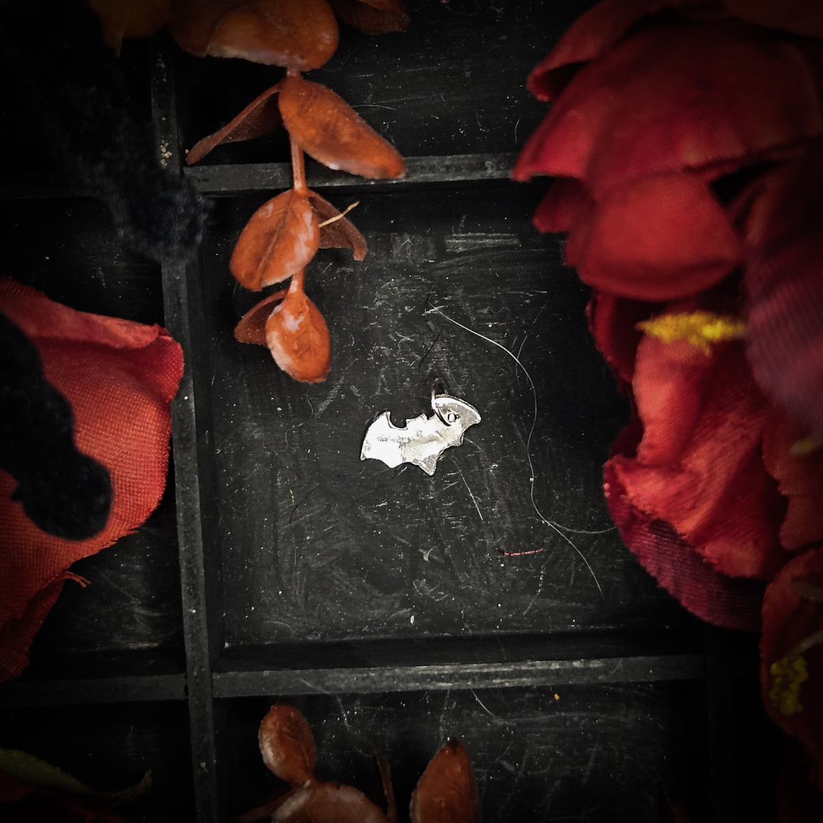 A mini sterling silver bat charm, displayed on a dark textured background surrounded by red and orange autumn leaves and flowers.