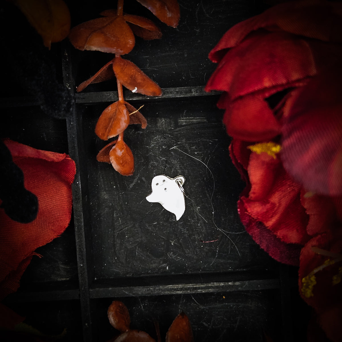 A small sterling silver flying ghost charm, displayed on a dark textured background surrounded by red and orange autumn leaves and flowers.