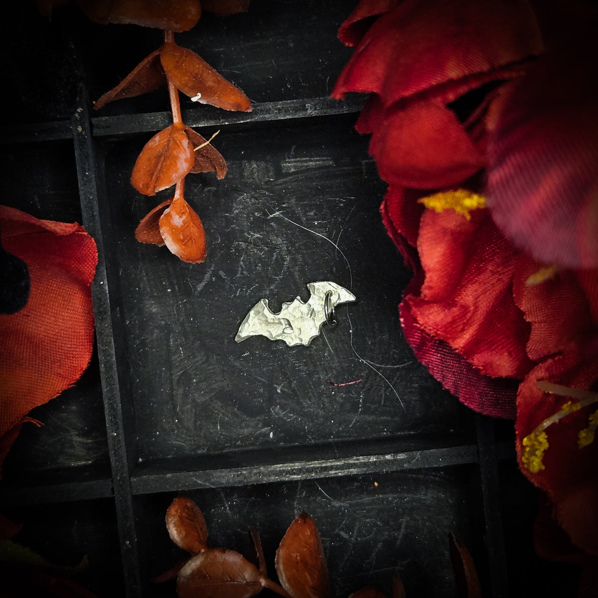 A small sterling silver bat charm, displayed on a dark textured background surrounded by red and orange autumn leaves and flowers.