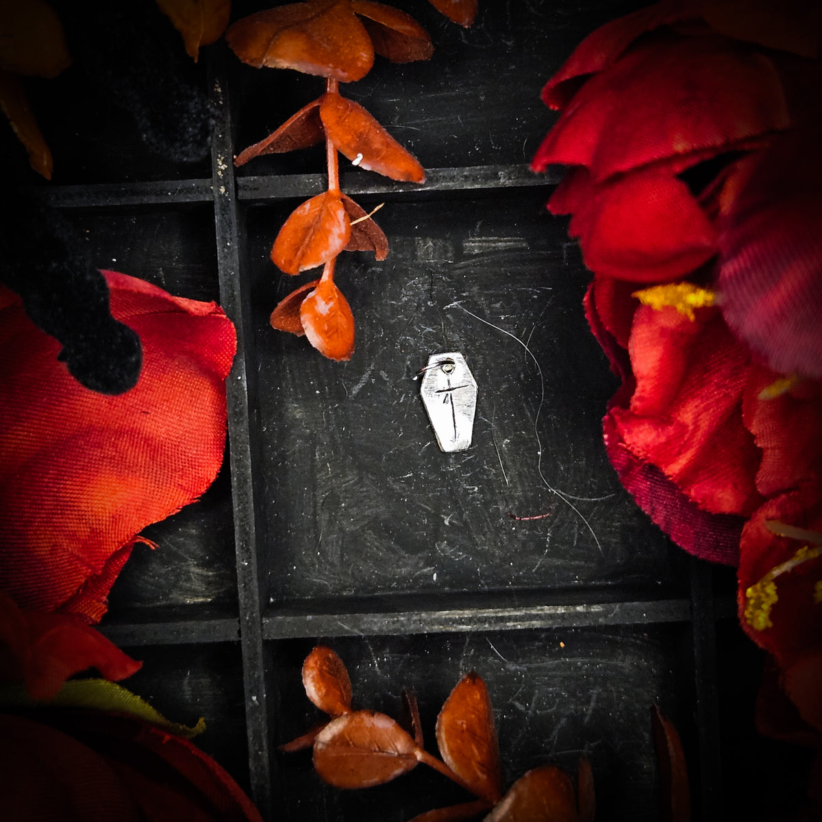 A small sterling silver coffin with cross charm, displayed on a dark textured background surrounded by red and orange autumn leaves and flowers.