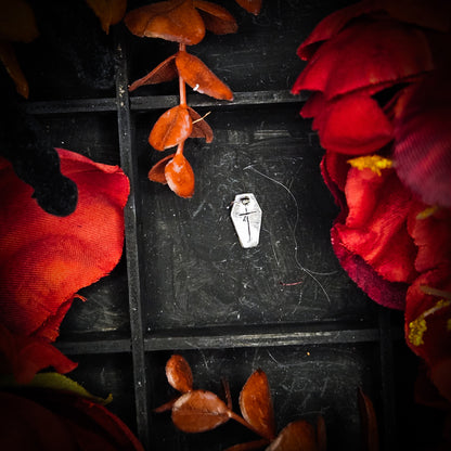 A small sterling silver coffin with cross charm, displayed on a dark textured background surrounded by red and orange autumn leaves and flowers.