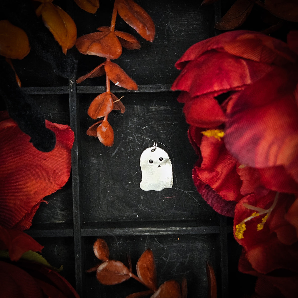 A small sterling silver ghost-shaped pendant with black eyes against a dark, decorative background framed by red and orange fabric flowers.
