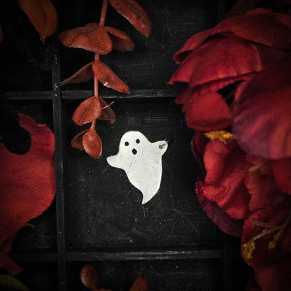 A large sterling silver ghost-shaped pendant with black eyes and mouth details, displayed on a dark textured background surrounded by red fabric flowers and dried red leaves.