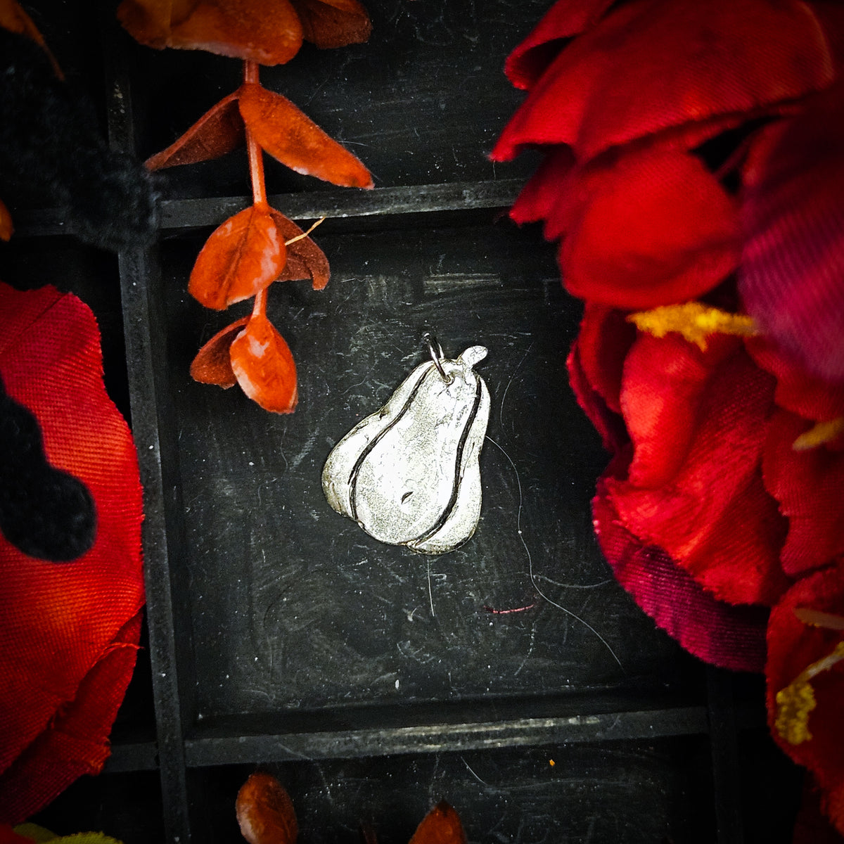 A sterling silver pumpkin-shaped pendant with a textured, hammered finish, displayed on a dark wooden background surrounded by red and orange fabric flowers.