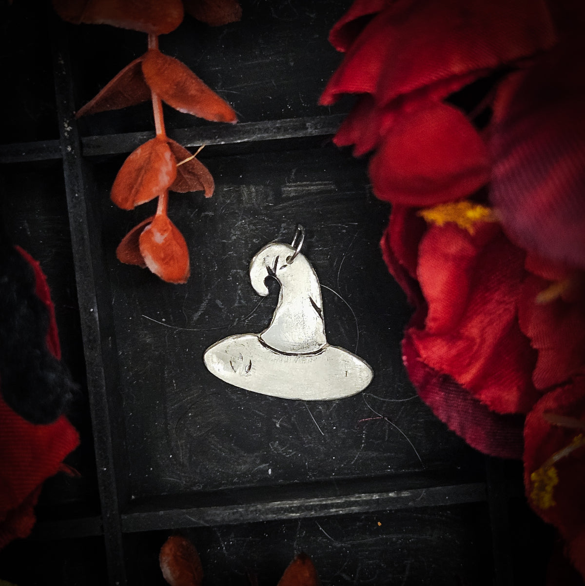 A large sterling silver witches hat pendant, displayed on a dark textured background surrounded by red and orange autumn leaves and flowers.