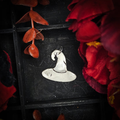 A large sterling silver witches hat pendant, displayed on a dark textured background surrounded by red and orange autumn leaves and flowers.