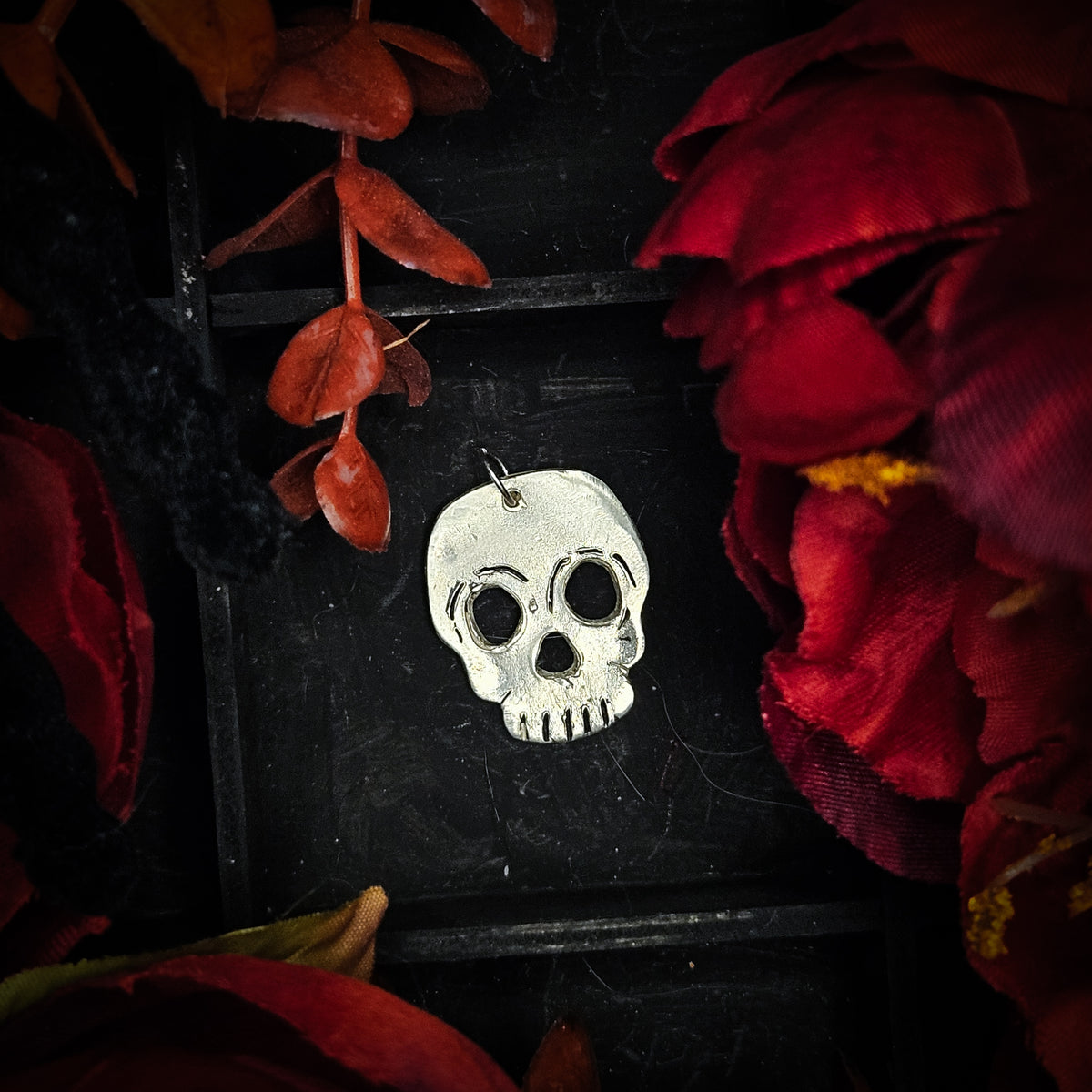 A silver skull pendant with a blackened, oxidized finish, displayed on a black background decorated with red dried flowers and leaves.