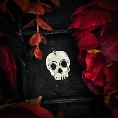 A silver skull pendant with a blackened, oxidized finish, displayed on a black background decorated with red dried flowers and leaves.
