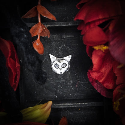 A small sterling silver cat-shaped pendant or charm with engraved eyes and nose, centered on a dark textured background surrounded by red and orange dried flowers.