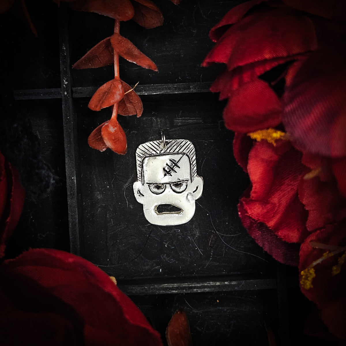 A sterling silver pendant shaped like Frankenstein's monster head with black line details, displayed against a dark background framed by red flowers and leaves.