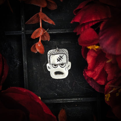 A sterling silver pendant shaped like Frankenstein's monster head with black line details, displayed against a dark background framed by red flowers and leaves.