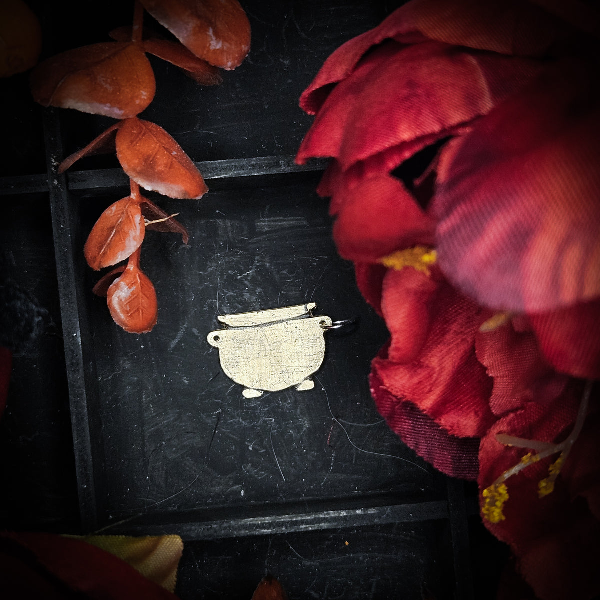A large brass charm shaped like a cauldron, displayed on a dark tray with dried orange leaves and red fabric flowers around it.