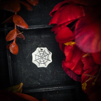 A sterling silver spider web pendant, displayed on a dark textured background surrounded by red and orange autumn leaves and flowers.