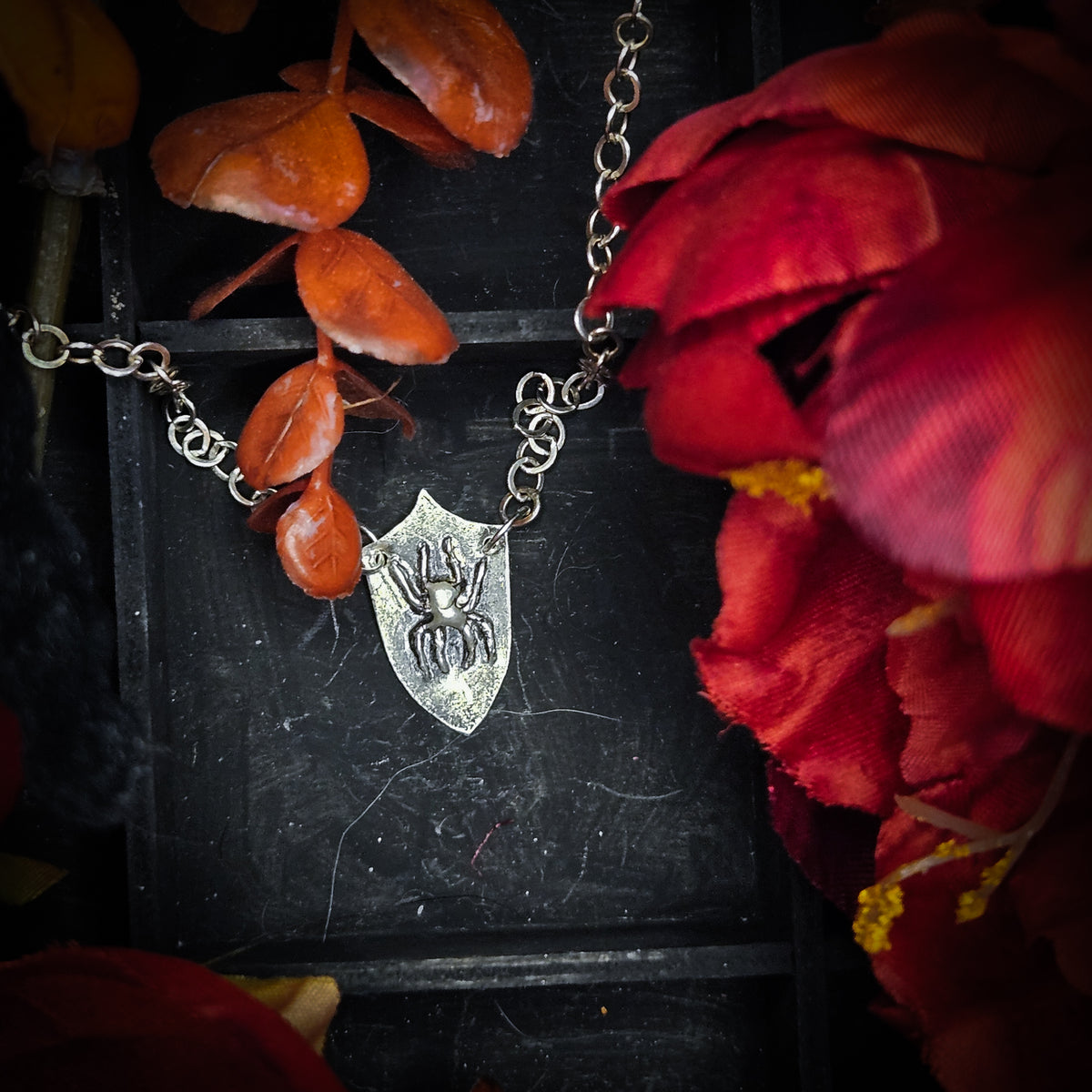 A silver-toned chain necklace with a shield-shaped pendant featuring an embossed spider, styled with dried orange leaves and red fabric flowers on a dark textured background.