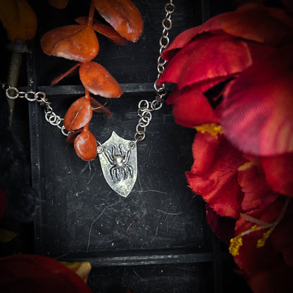 A silver-toned chain necklace with a shield-shaped pendant featuring an embossed spider, styled with dried orange leaves and red fabric flowers on a dark textured background.