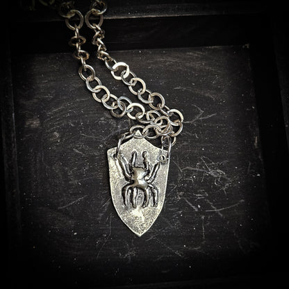 A silver-toned shield pendant necklace featuring an embossed spider design, hanging from a link chain on a dark textured background.