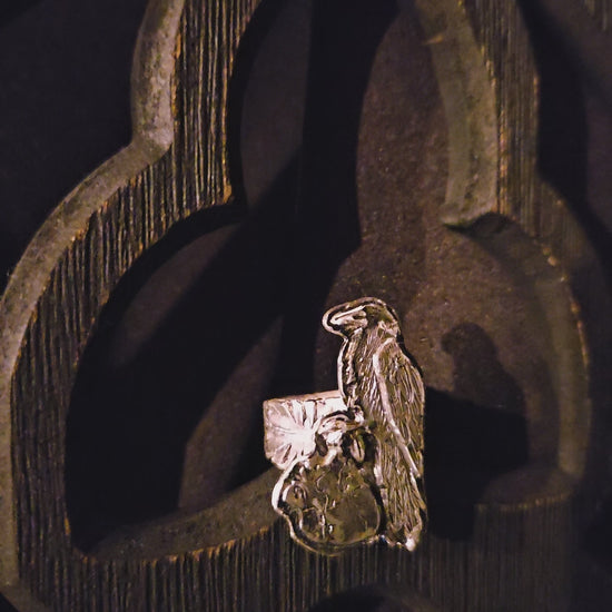 Sterling silver raven and human heart ring with a feather band on a dark wood background